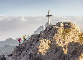 Two mountaineers climb a rocky ridge in the Dolomites, approaching a striking summit cross with dramatic peaks rising through the clouds in the background.