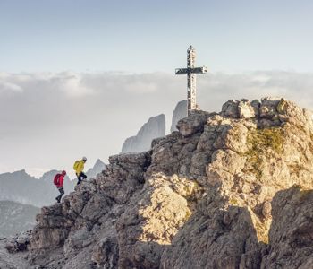 Zwei Bergsteiger erklimmen einen felsigen Grat in den Dolomiten und nähern sich einem markanten Gipfelkreuz, während im Hintergrund markante Bergspitzen aus den Wolken ragen.