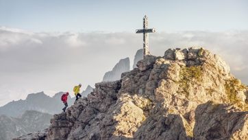Two mountaineers climb a rocky ridge in the Dolomites, approaching a striking summit cross with dramatic peaks rising through the clouds in the background.