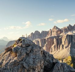 Zwei Wanderer stehen jubelnd neben einem Gipfelkreuz auf einem felsigen Berggipfel in den Dolomiten, umgeben von beeindruckenden Felsformationen und weitem Panorama unter blauem Himmel.