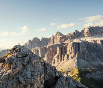 Zwei Wanderer stehen jubelnd neben einem Gipfelkreuz auf einem felsigen Berggipfel in den Dolomiten, umgeben von beeindruckenden Felsformationen und weitem Panorama unter blauem Himmel.