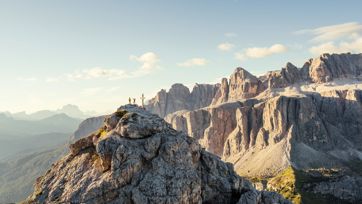 Due escursionisti esultano accanto a una croce di vetta su una cima rocciosa nelle Dolomiti, circondati da imponenti formazioni rocciose e un ampio panorama sotto un cielo azzurro.