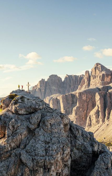 Zwei Wanderer stehen jubelnd neben einem Gipfelkreuz auf einem felsigen Berggipfel in den Dolomiten, umgeben von beeindruckenden Felsformationen und weitem Panorama unter blauem Himmel.