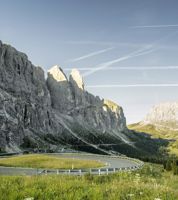 Panoramablick auf die kurvenreiche Passstraße unterhalb des imposanten Sellamassivs in den Dolomiten, eingebettet in eine grüne, sommerliche Alpenlandschaft.