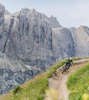 Ein Mountainbiker fährt eine kurvenreiche Schotterpiste entlang, flankiert von grünen Wiesen mit Wildblumen, im Hintergrund die imposanten Felswände der Dolomiten.