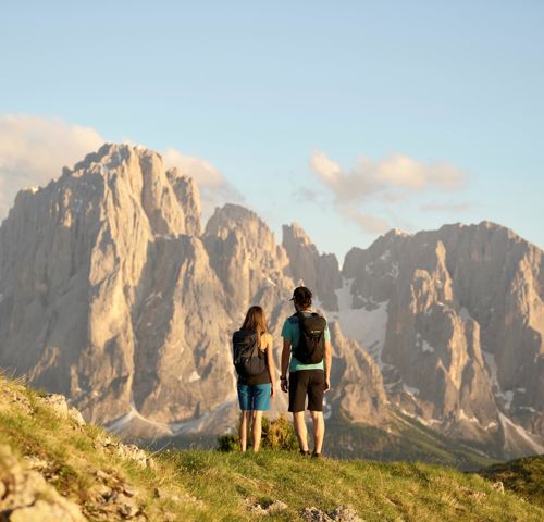 Ein Mann und eine Frau wandern entlang einer Almwiese, im Hntergrund die Dolomiten