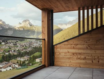 Moderner Balkon mit Glasgeländer und Holzelementen, Blick auf das Dorf Wolkenstein und die Dolomiten.