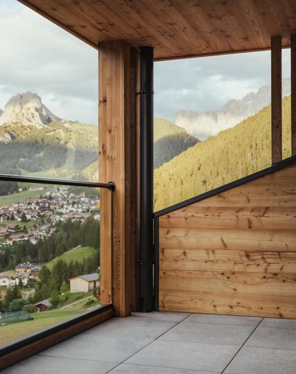 Modern balcony with glass railing and wooden elements, overlooking Selva di Val Gardena and the Dolomites.