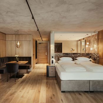 Junior Suite Exclusive mit Balkon, gemütlichem Doppelbett, moderner Holzverkleidung, stilvoller Sitzecke und offenem Badezimmer mit Marmor-Details.