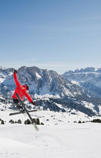 Ein Skifahrer führt einen Sprung in der Luft aus, mit einer spektakulären Winterlandschaft der verschneiten Dolomiten im Hintergrund.