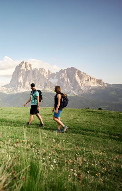 Ein Mann und eine Frau wandern entlang einer Almwiese, im Hntergrund die Dolomiten